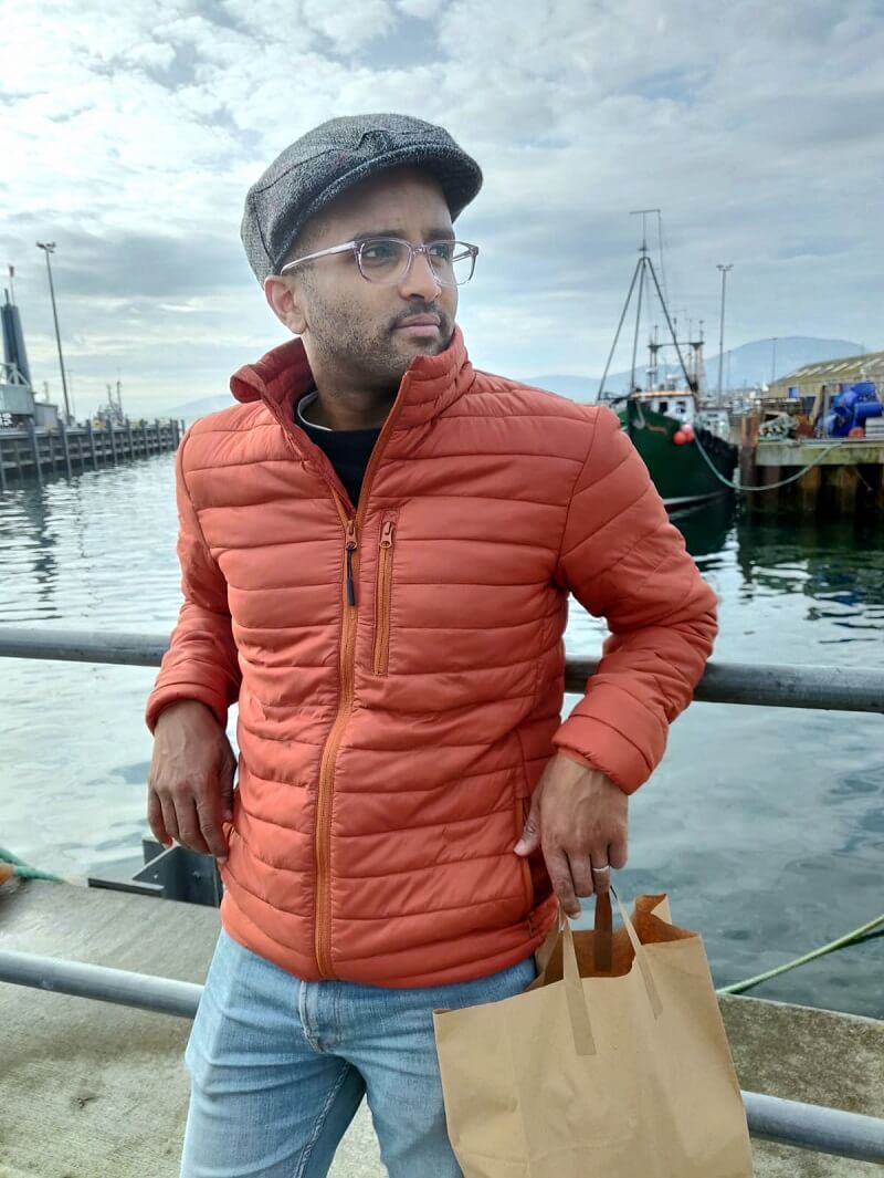 Ahmed Khalifa, a black man wearing a flat cap and an orange puffy coat, leaning against the rails with the view of the sea and the harbour in the background.