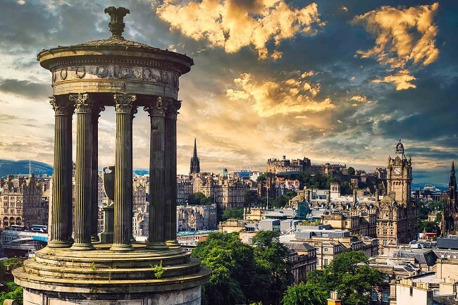 The skyline of the city centre of Edinburth at the top of Calton Hill which has pillared stones. In the backdrop, there are various buildings of including a tall Victorian style building with a clock at the top and the Edinburgh castle to the left of it.