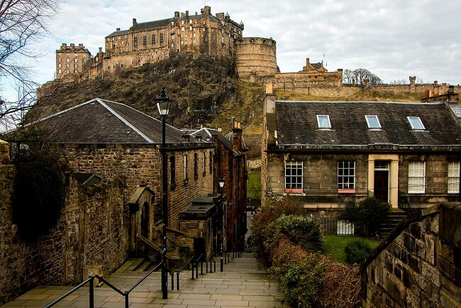 A side street of Edinburgh with steps, old-fashioned street lantern, stoned buildings and the Edinburgh Castle in clear view looming over it